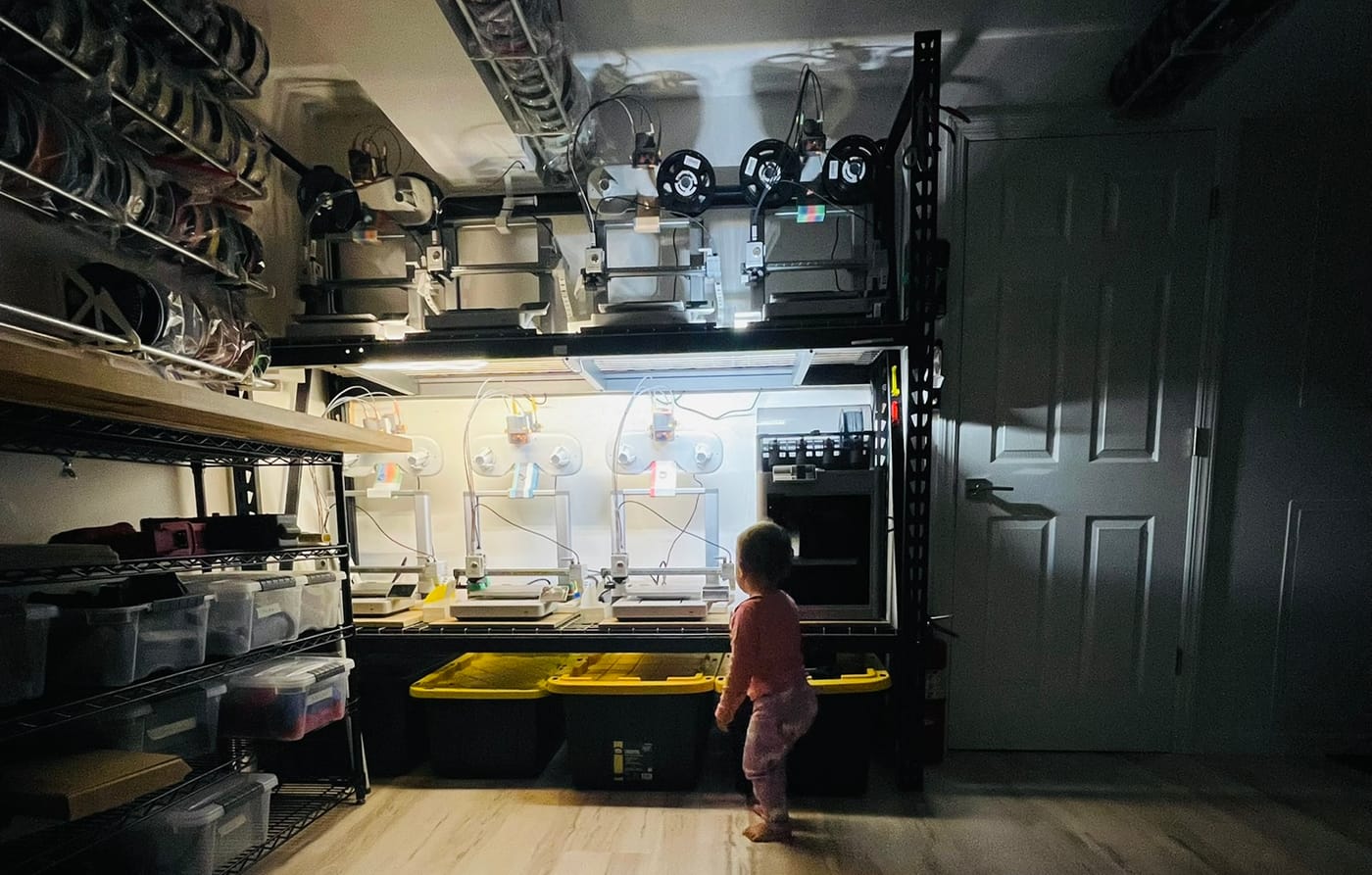 The first wall of 3D printers glowing in a basement workshop, with a toddler standing in front watching them run